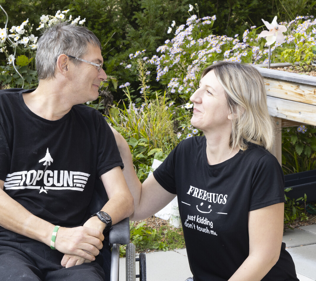 Im Au&szlig;enbereich des Wohnverbunds K&auml;the Dorsch hockt eine BALANCE Mitarbeiterin neben einem Bewohner im Rollstuhl. Sie sehen sich an.