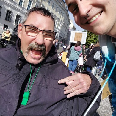 Ein Bewohner und ein Mitarbeiter von BALANCE machen ein fr&ouml;hliches Selfie auf einer Demonstration f&uuml;r Inklusion in Wien. Im Vordergrund lacht ein Mann mit Brille und Schnurrbart ausgelassen in die Kamera, tr&auml;gt eine gr&uuml;ne Trillerpfeife um den Hals und zeigt mit der Hand ein Rock-Geste-Symbol. Rechts im Bild l&auml;chelt ein j&uuml;ngerer Mitarbeiter mit Piercings herzlich. Im Hintergrund sind weitere Teilnehmer der Demonstration, Transparente und historische Geb&auml;ude unter blauem Himmel zu sehen.