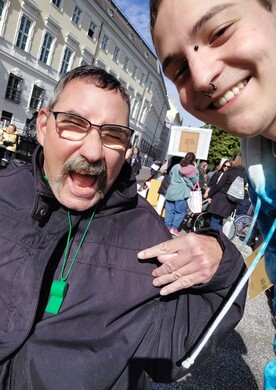 Ein Bewohner und ein Mitarbeiter von BALANCE machen ein fr&ouml;hliches Selfie auf einer Demonstration f&uuml;r Inklusion in Wien. Im Vordergrund lacht ein Mann mit Brille und Schnurrbart ausgelassen in die Kamera, tr&auml;gt eine gr&uuml;ne Trillerpfeife um den Hals und zeigt mit der Hand ein Rock-Geste-Symbol. Rechts im Bild l&auml;chelt ein j&uuml;ngerer Mitarbeiter mit Piercings herzlich. Im Hintergrund sind weitere Teilnehmer der Demonstration, Transparente und historische Geb&auml;ude unter blauem Himmel zu sehen.