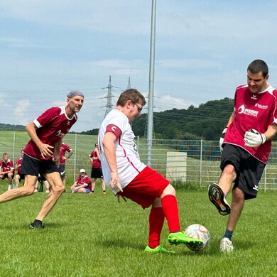 Drei M&auml;nner spielen leidenschaftlich Fu&szlig;ball auf einem gr&uuml;nen Rasenplatz unter blauem Himmel. Im Vordergrund kickt ein Spieler in wei&szlig;em Trikot und roten Shorts den Ball, w&auml;hrend ein Gegenspieler in rotem Trikot und schwarzen Shorts versucht, ihn zu blocken. Ein dritter Spieler in Rot l&auml;uft im Hintergrund mit. Im unscharfen Hintergrund sind weitere Personen am Spielfeldrand sowie bewaldete H&uuml;gel und Strommasten zu sehen.