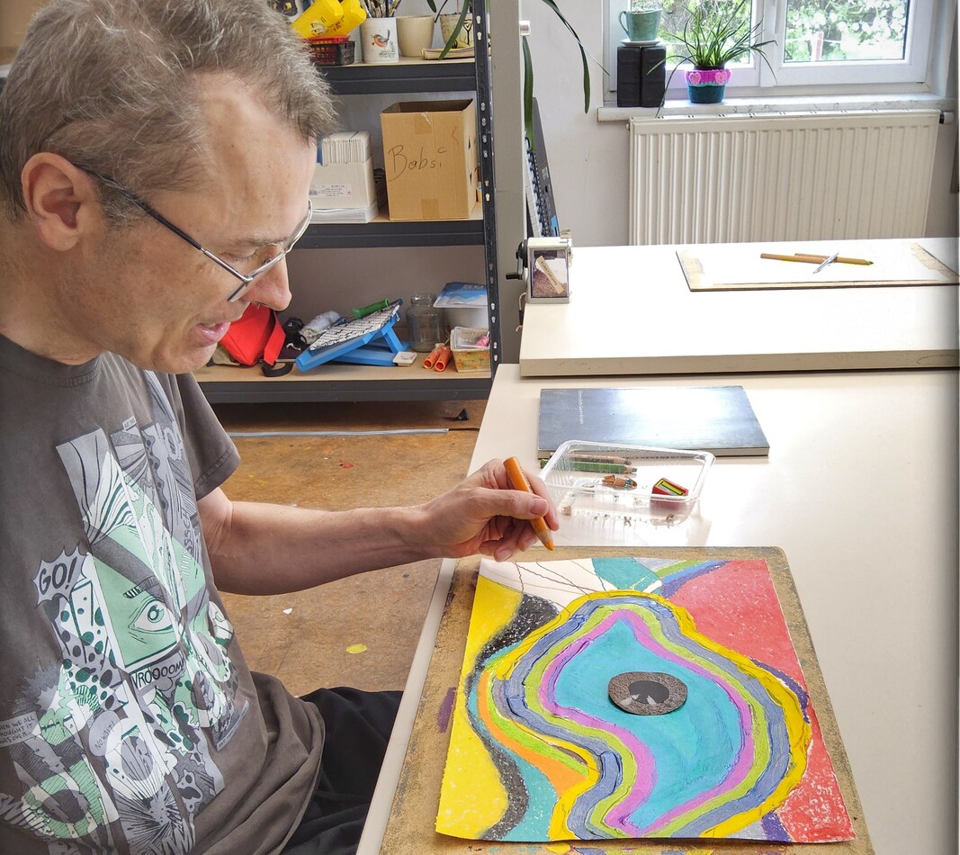 Der K&uuml;nstler Erwin Hasitzka arbeitet konzentriert in seinem Atelier in Maria Ponsee. Er sitzt an einem gro&szlig;en Arbeitstisch und zeichnet mit einem orangefarbenen Stift an einem abstrakten, farbenfrohen Werk. Das Bild vor ihm besteht aus organischen, wellenf&ouml;rmigen Linien in leuchtenden Farben wie T&uuml;rkis, Pink, Gelb und Rot. Im Vordergrund liegen verschiedene Zeichenutensilien bereit, und im Hintergrund ist das helle, mit Regalen und Materialien ausgestattete Atelier zu sehen.