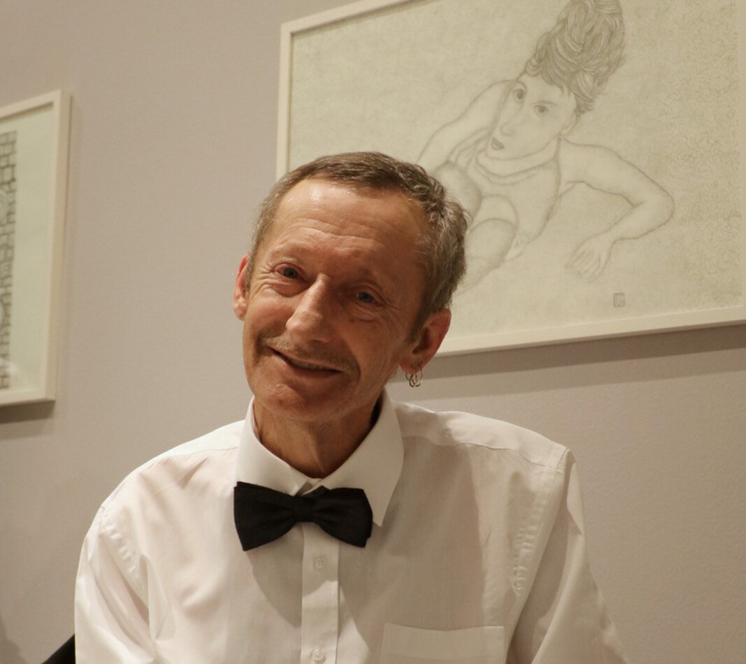 Ein Portr&auml;t des K&uuml;nstlers Werner Roitner bei einer Ausstellung. Er sitzt l&auml;chelnd in seinem Rollstuhl und tr&auml;gt ein elegantes wei&szlig;es Hemd mit einer schwarzen Fliege. Im Hintergrund ist eines seiner Werke zu sehen: Eine gerahmte, feine Bleistiftzeichnung auf hellem Grund, die eine stilisierte menschliche Figur in einer dynamischen Pose zeigt. Die Szene vermittelt eine festliche und stolze Stimmung.