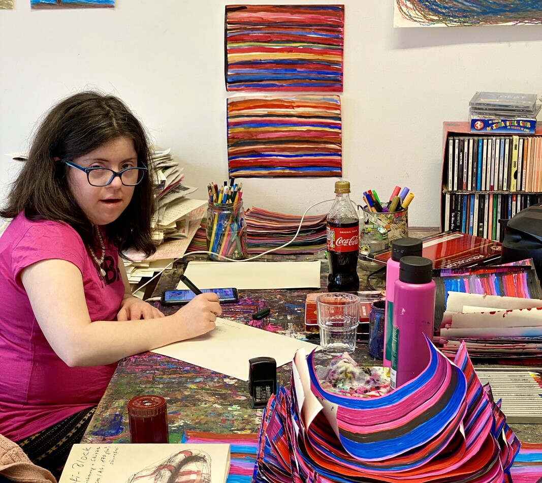 Die K&uuml;nstlerin Andrea Meija Rocha sitzt an ihrem Arbeitsplatz, sie h&auml;lt einen Stift in der Hand, auf dem Tisch befinden sich verschiedene Kunstwerke, und Arbeitsmaterialien im Hintergrund an der Wand h&auml;ngen Bilder