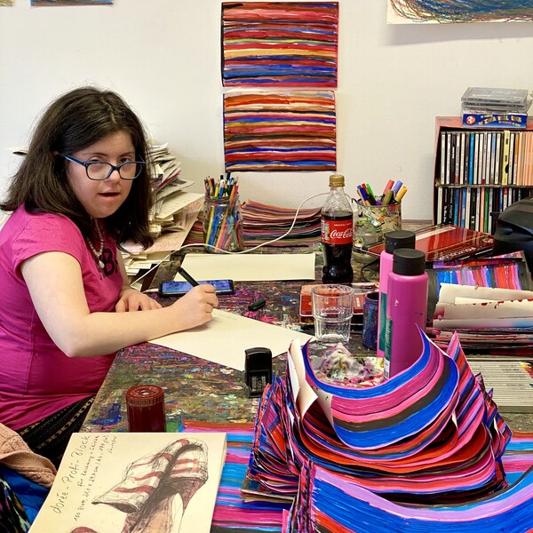 Die K&uuml;nstlerin Andrea Meija Rocha sitzt an ihrem Arbeitsplatz, sie h&auml;lt einen Stift in der Hand, auf dem Tisch befinden sich verschiedene Kunstwerke, und Arbeitsmaterialien im Hintergrund an der Wand h&auml;ngen Bilder