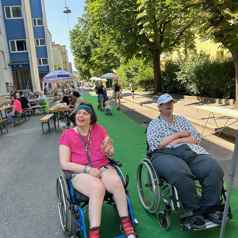 Zwei Bewohner im Rollstuhl lachen ausgelassen beim Gr&auml;tzlfest in der K&auml;the-Dorsch-Gasse. Eine Frau im pinken T-Shirt und ein Mann im gemusterten Hemd mit wei&szlig;er Kappe sitzen auf einem langen gr&uuml;nen Teppich, der mitten auf der Stra&szlig;e ausgerollt ist. Im Hintergrund der sonnigen, baumbestandenen Gasse sind Festb&auml;nke, Sonnenschirme und weitere Besucher zu sehen, die das Nachbarschaftsfest genie&szlig;en.