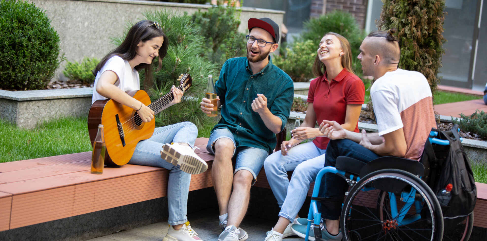 Eine Gruppe junger Freunde, darunter ein junger Mann mit Rolstuhl sitzt lachend im Freien; eine Frau spielt Gitarre, w&auml;hrend die anderen singen und Getr&auml;nke halten.