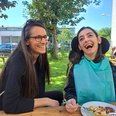 Zwei Frauen lachen ausgelassen an einem Holztisch beim Sommerfest der Tagesstruktur Soho. Eine Frau mit langen dunklen Haaren und Brille blickt freudig zu ihrer Begleiterin, die in einem Rollstuhl sitzt, ein t&uuml;rkisfarbenes L&auml;tzchen tr&auml;gt und den Kopf mit weit offenem Mund vor Lachen nach hinten wirft. Vor ihnen steht ein Pappteller mit einem St&uuml;ck Torte. Die Szene spielt im Freien auf einer gr&uuml;nen Wiese unter schattigen B&auml;umen bei strahlendem Sonnenschein