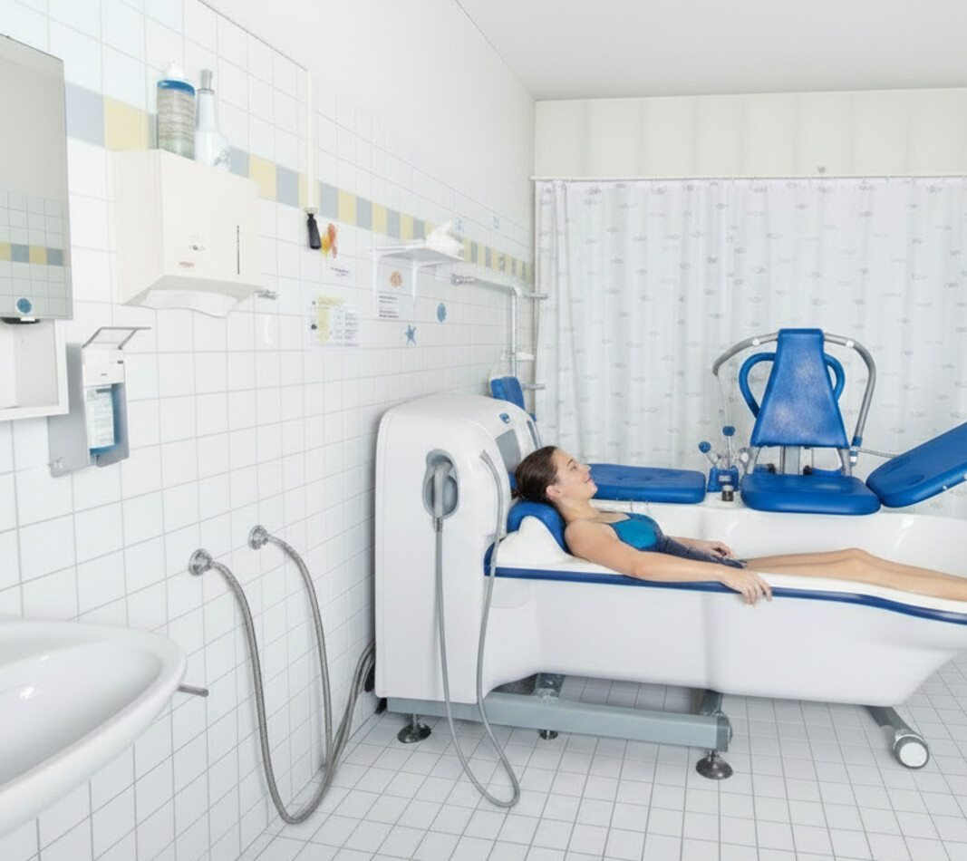 Eine Person liegt entspannt in einer wei&szlig;en Pflegebadewanne in einem hell gefliesten Therapieraum. Die Wanne ist mit einem blauen Patientenlifter und spezialisierten Armaturen ausgestattet. Im Hintergrund sind wei&szlig;e Wandfliesen, ein Waschbecken und medizinische Spender zu sehen.