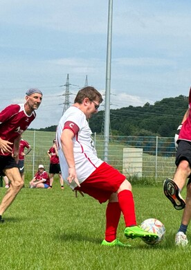 Drei M&auml;nner spielen leidenschaftlich Fu&szlig;ball auf einem gr&uuml;nen Rasenplatz unter blauem Himmel. Im Vordergrund kickt ein Spieler in wei&szlig;em Trikot und roten Shorts den Ball, w&auml;hrend ein Gegenspieler in rotem Trikot und schwarzen Shorts versucht, ihn zu blocken. Ein dritter Spieler in Rot l&auml;uft im Hintergrund mit. Im unscharfen Hintergrund sind weitere Personen am Spielfeldrand sowie bewaldete H&uuml;gel und Strommasten zu sehen.
