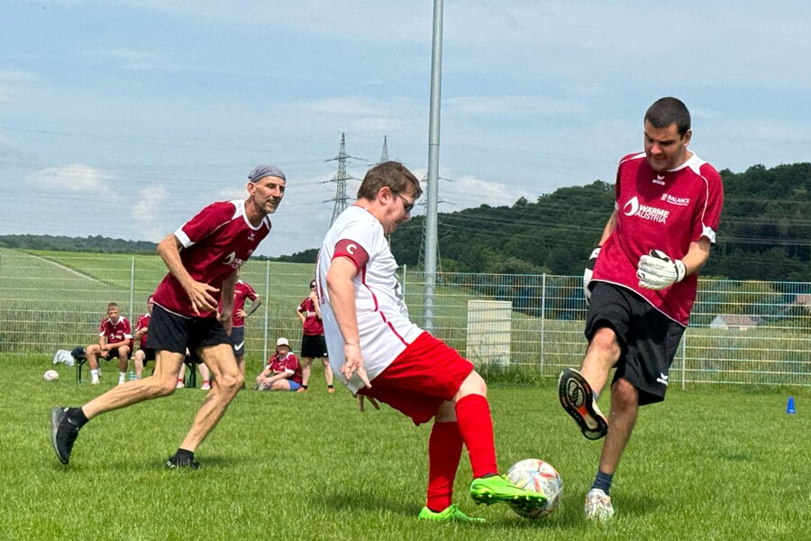 Drei Personen mit und ohne Behinderung spielen gemeinsam Fu&szlig;ball auf einer Wiese.