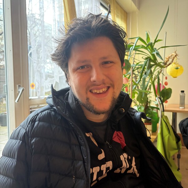 Ein l&auml;chelnder junger Mann mit dunklem Haar und schwarzer Steppjacke in einem hellen Raum mit Zimmerpflanzen.