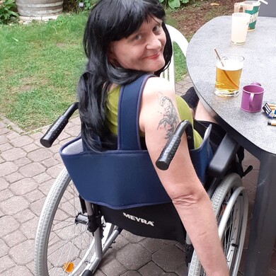 Eine Bewohnerin mit langen schwarzen Haaren und einem auff&auml;lligen Skorpion-Tattoo am Oberarm sitzt in ihrem Rollstuhl im Garten der Wohngemeinschaft B&ouml;ckh. Sie tr&auml;gt ein olivgr&uuml;nes Top, hat die Lippen rot geschminkt und blickt mit einem verschmitzten L&auml;cheln &uuml;ber die Schulter direkt in die Kamera. Sie befindet sich auf einer gepflasterten Terrasse an einem grauen Gartentisch, auf dem Gl&auml;ser und Getr&auml;nke stehen. Im Hintergrund ist eine gr&uuml;ne Gartenanlage mit Pflanzen und einem gro&szlig;en Holzfass zu sehen.