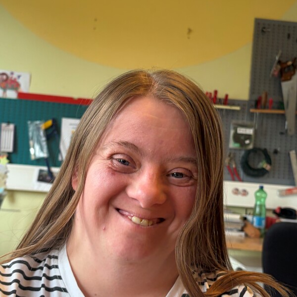 Portr&auml;taufnahme von Suzana, einer Frau mit langen, braunen Haaren und einem herzlichen L&auml;cheln. Sie tr&auml;gt ein schwarz-wei&szlig; gestreiftes T-Shirt. Der Hintergrund zeigt eine helle Werkstatt mit Werkzeugw&auml;nden und einer gelben Decke.