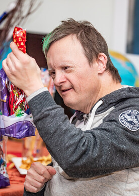 Ein Mann mit Trisomie 21 und einer markanten gr&uuml;nen Haarstr&auml;hne blickt l&auml;chelnd auf eine lila-rote Geschenkverpackung, die er in der Hand h&auml;lt. Er tr&auml;gt einen sportlichen, grau-melierten Kapuzenpullover mit einem runden Logo-Aufn&auml;her am &Auml;rmel. Vor ihm auf einem Tisch stehen weitere weihnachtlich verpackte Geschenkt&uuml;ten. Der Hintergrund ist hell und leicht unscharf.