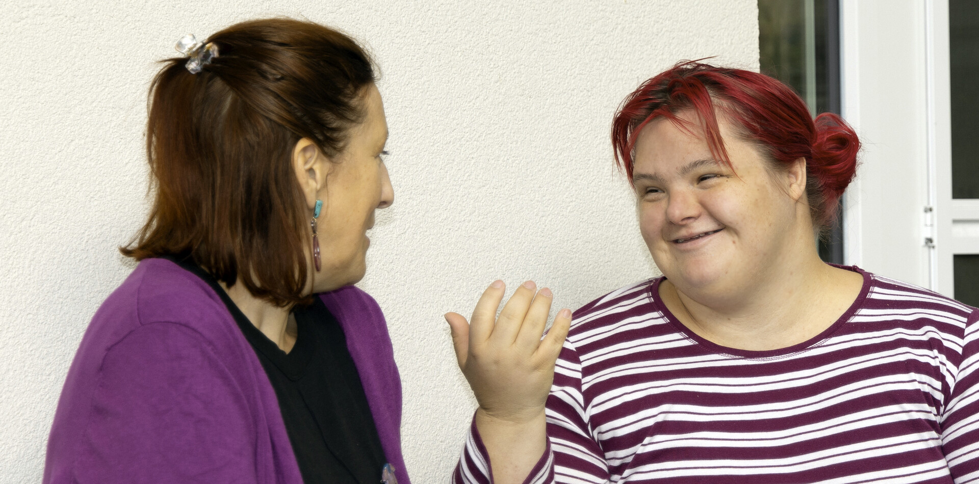 Eine Frau mit braunen Haaren und lila Weste und eine Frau mit roten Haaren, die ein lila-wei&szlig; gestreiftes Oberteil tr&auml;gt sind in einem Gespr&auml;ch. Beide l&auml;cheln und wirken fr&ouml;hlich und entspannt.