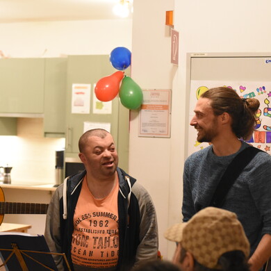 Ein Bewohner und ein Unterst&uuml;tzer stehen nebeneinander und lachen sich an. Im Hintergrund sieht man drei Luftballons an der Wand h&auml;ngen. Links sieht man ein kleines St&uuml;ck einer Gitarre.