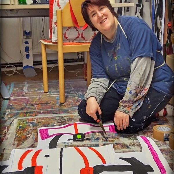 Die K&uuml;nstlerin Barbara Plak kniet am Boden, h&auml;lt eine Schere in der Hand und lacht in die Kamera, auf dem Boden vor ihr sieht man ein gro&szlig;es Kunstwerk aus Papier von dem ein Teil herausgeschnitten wurde.