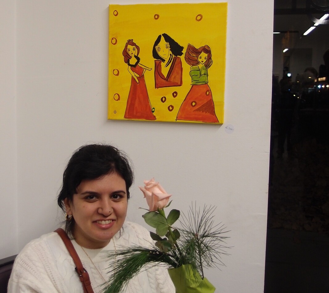 Die K&uuml;nstlerin Juka Abdilwahid steht in einer Ausstellung vor einem ihrer Bilder und l&auml;chelt in die Kamera. rechts von ihr ist eine hellrosa Rose. Das Kunstwerk zeit drei Frauengestalten in roten Kleidern vor gelbem Hintergrund.