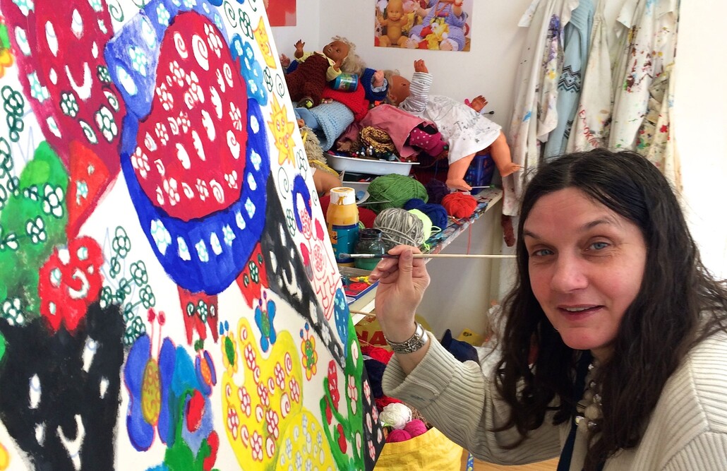 Eine Frau mit dunklen langen Haaren h&auml;lt einen Pinsel in der Hand und malt an einem gro&szlig;en bunten Bild. Sie schaut in die Kamera.