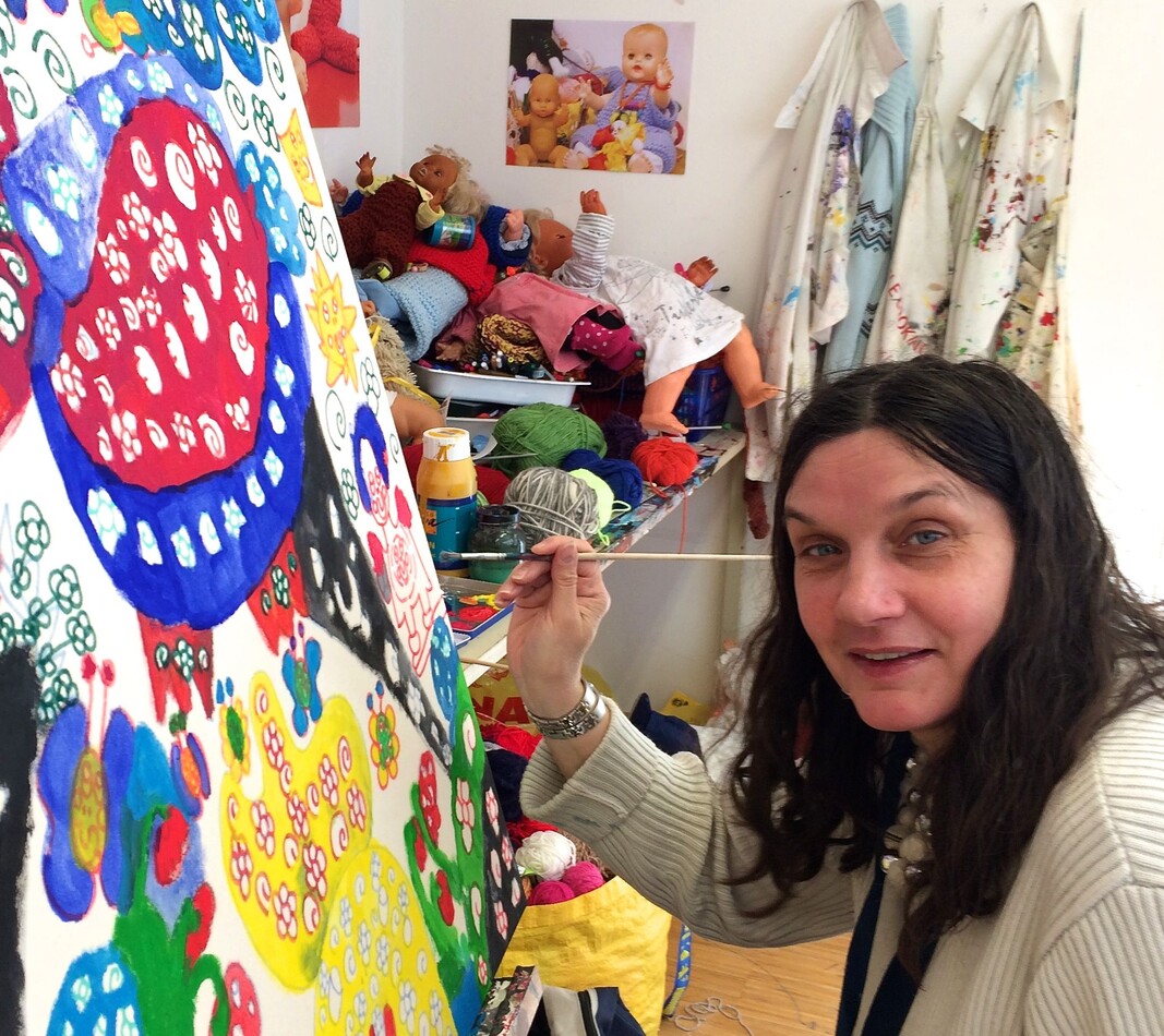 Die K&uuml;nstlerin Steffi Wimmer arbeitet im Atelier bildBalance in Wien an einem gro&szlig;formatigen, farbenfrohen Gem&auml;lde. Mit einem Pinsel f&uuml;gt sie einem Bild, das aus vielen bunten Mustern, Spiralen und floralen Elementen besteht, Details hinzu. Sie hat lange dunkle Haare, tr&auml;gt einen hellen Strickpullover und blickt l&auml;chelnd zur Seite. Im Hintergrund des Ateliers sind Regale mit Wolle und Puppen sowie Malkittel zu sehen, die an der Wand h&auml;ngen.