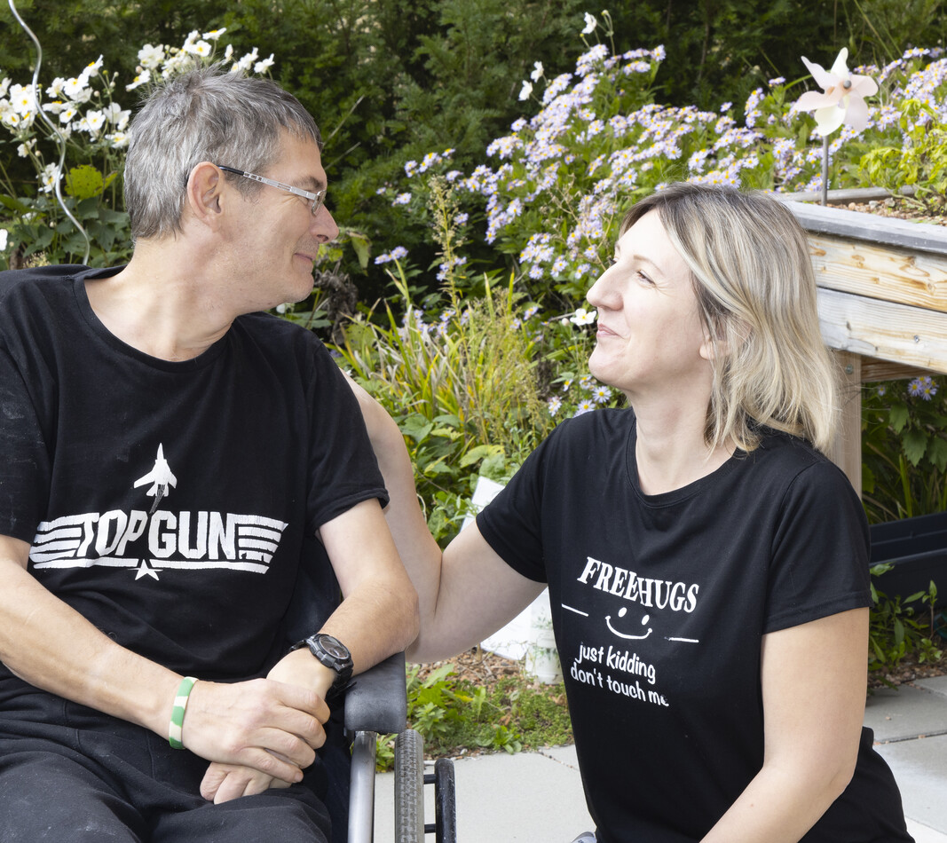 Ein l&auml;chelnder Mann im Rollstuhl und eine Frau knien im Garten nebeneinander und schauen sich freundlich an. Der Mann tr&auml;gt ein schwarzes T-Shirt mit dem &bdquo;Top Gun&ldquo;-Logo, die Frau ein schwarzes T-Shirt mit der Aufschrift &bdquo;FREE HUGS &ndash; just kidding, don't touch me&ldquo;. Im Hintergrund sind bl&uuml;hende Gartenpflanzen und ein Hochbeet zu sehen.