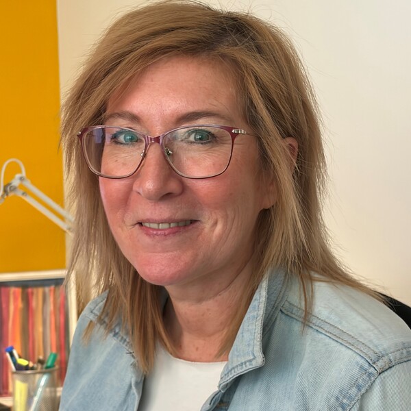 Mitarbeiterin Cordula l&auml;chelt freundlich in die Kamera. Sie tr&auml;gt eine markante Brille mit dunklem Rahmen, eine helle Jeansjacke und hat mittellanges, blondes Haar.