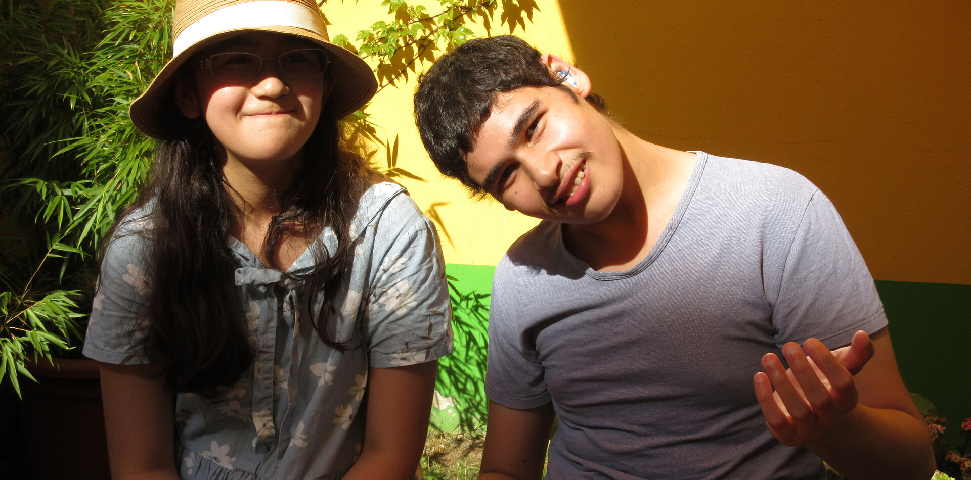 Eine junge Frau mit Sonnenhut und T-Shirt und ein junger Mann im T-Shirt sitzen im Hof der WG Salzach und l&auml;cheln in die Kamera
