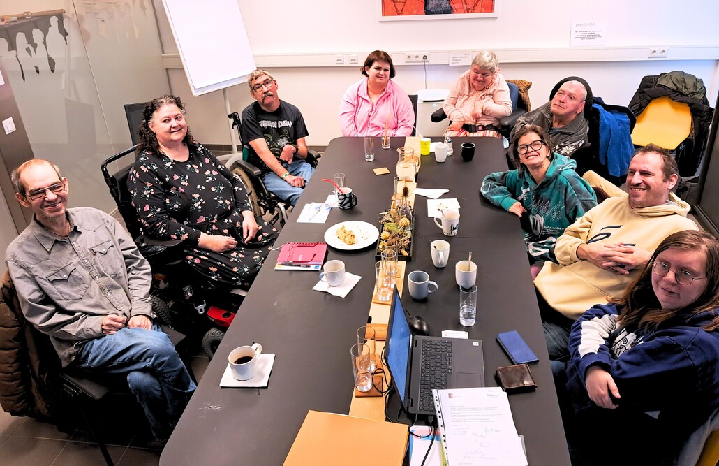 Das Wicky Team &ndash; die Ideen- und Beschwerdestelle f&uuml;r Nutzer:innen &ndash; bei einer gemeinsamen Besprechung an einem gro&szlig;en, dunklen Konferenztisch in der BALANCE Zentrale. Neun Personen sitzen in einer gem&uuml;tlichen Runde zusammen, einige davon in Rollst&uuml;hlen. Auf dem Tisch stehen Kaffeetassen, Gl&auml;ser, ein Laptop und Unterlagen. Die Atmosph&auml;re ist konzentriert und freundlich. An der wei&szlig;en Wand im Hintergrund h&auml;ngen ein Flipchart und ein farbenfrohes, abstraktes Kunstwerk.