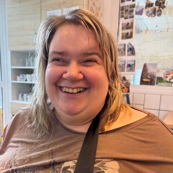 Portr&auml;tfoto von Melanie, einer herzlich lachenden Frau mit hellem, schulterlangem Haar. Sie tr&auml;gt ein braunes Oberteil mit floralen Mustern und einen schwarzen Umh&auml;ngegurt. Der Hintergrund zeigt einen hellen, freundlichen Raum mit Regalen und Fotos an der Wand.