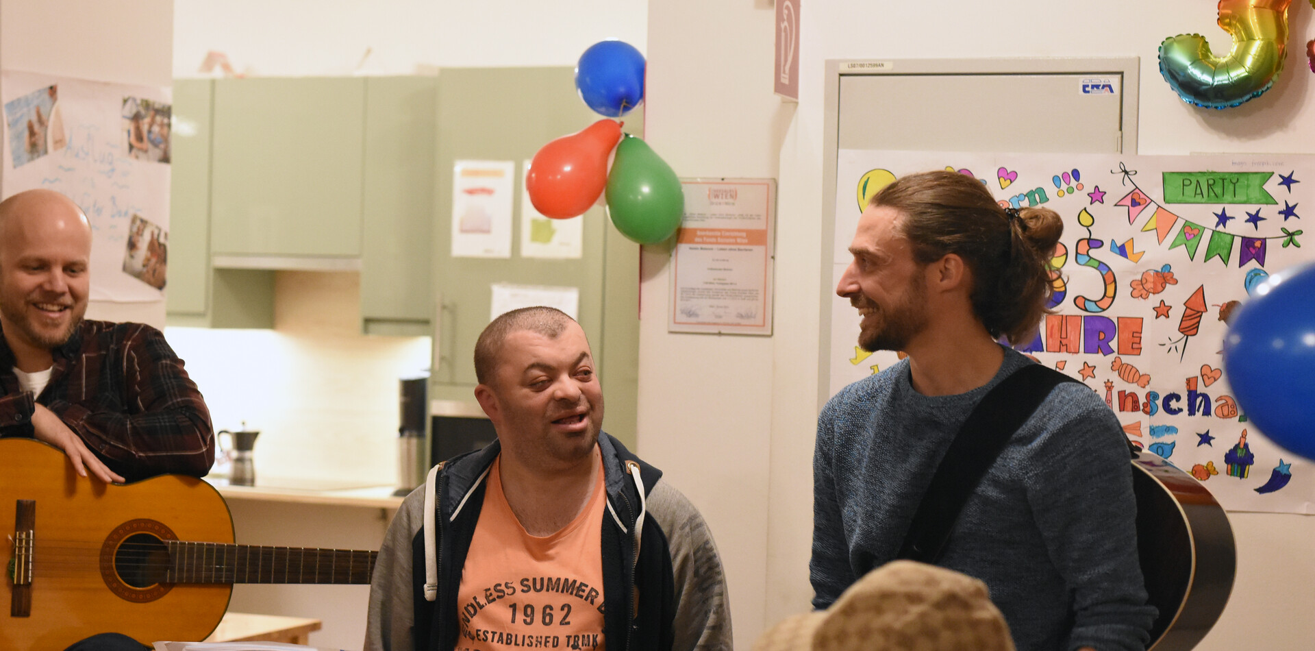 Ein Bewohner und ein Unterst&uuml;tzer stehen nebeneinander und lachen sich an. Im Hintergrund sieht man drei Luftballons an der Wand h&auml;ngen. Links sieht man ein kleines St&uuml;ck einer Gitarre.
