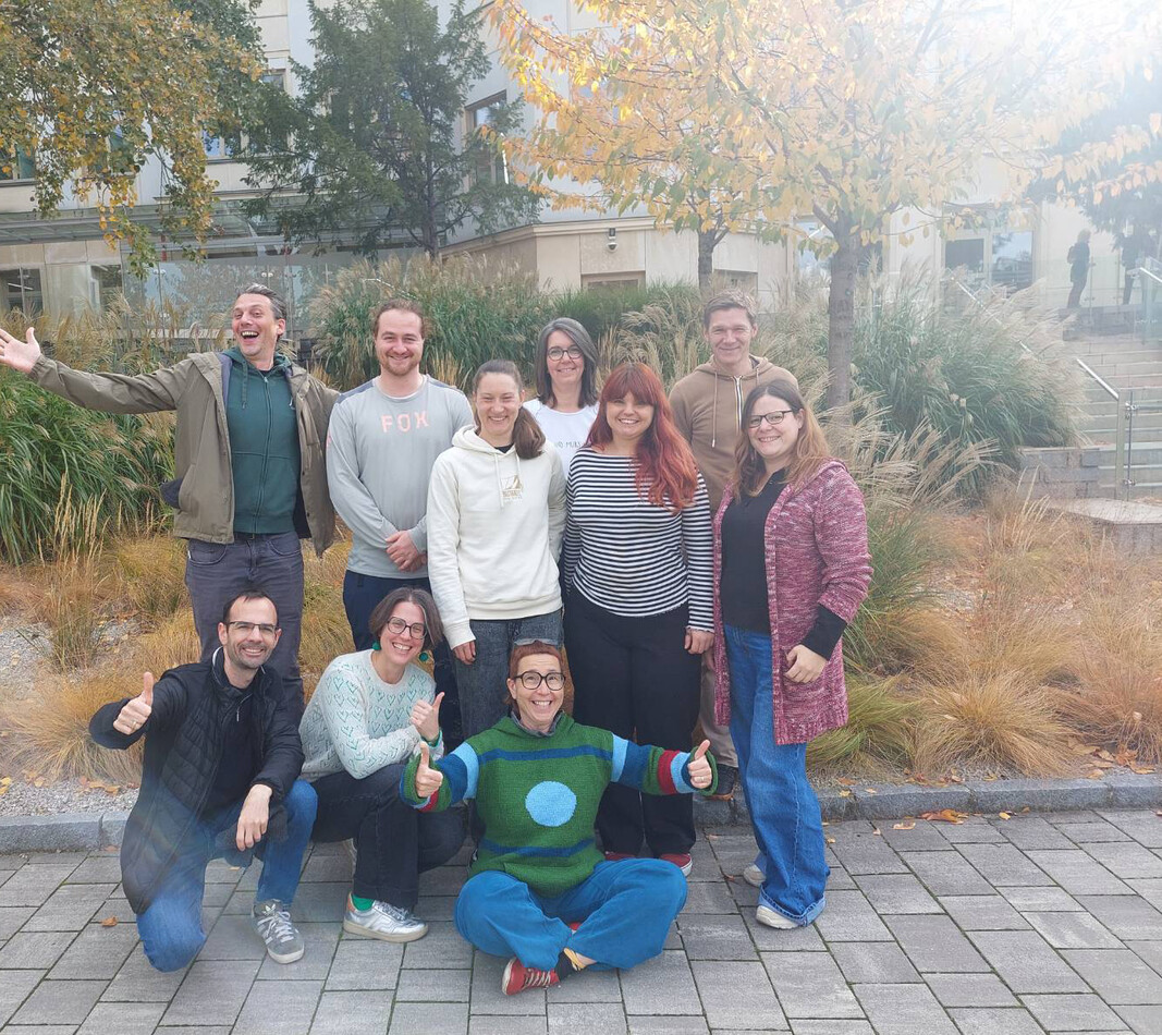 Ein fr&ouml;hliches Gruppenbild von zehn Betriebsr&auml;ten der Organisation BALANCE im Freien. Neun Personen stehen oder hocken in zwei Reihen vor einer herbstlich gestalteten Gr&uuml;nanlage mit hohen Ziergr&auml;sern und B&auml;umen, w&auml;hrend eine Person im Schneidersitz ganz vorne in der Mitte sitzt und beide Daumen hochstreckt. Alle l&auml;cheln oder lachen ausgelassen in die Kamera, ein Mann am linken Rand breitet jubelnd die Arme aus. Im Hintergrund ist die Fassade eines modernen Geb&auml;udes zu sehen.