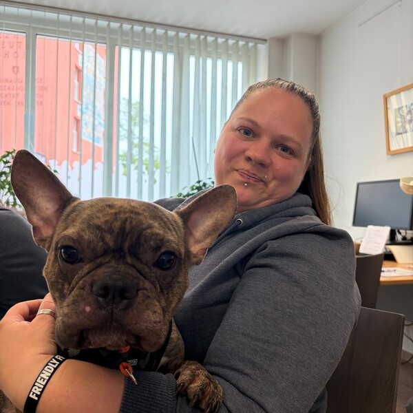 Dani l&auml;chelt in die Kamera und h&auml;lt Muffin, eine braun-schwarz gestromte Franz&ouml;sische Bulldogge, auf dem Scho&szlig;. Sie befinden sich in einem hellen B&uuml;ro. Dani tr&auml;gt einen grauen Kapuzenpullover.