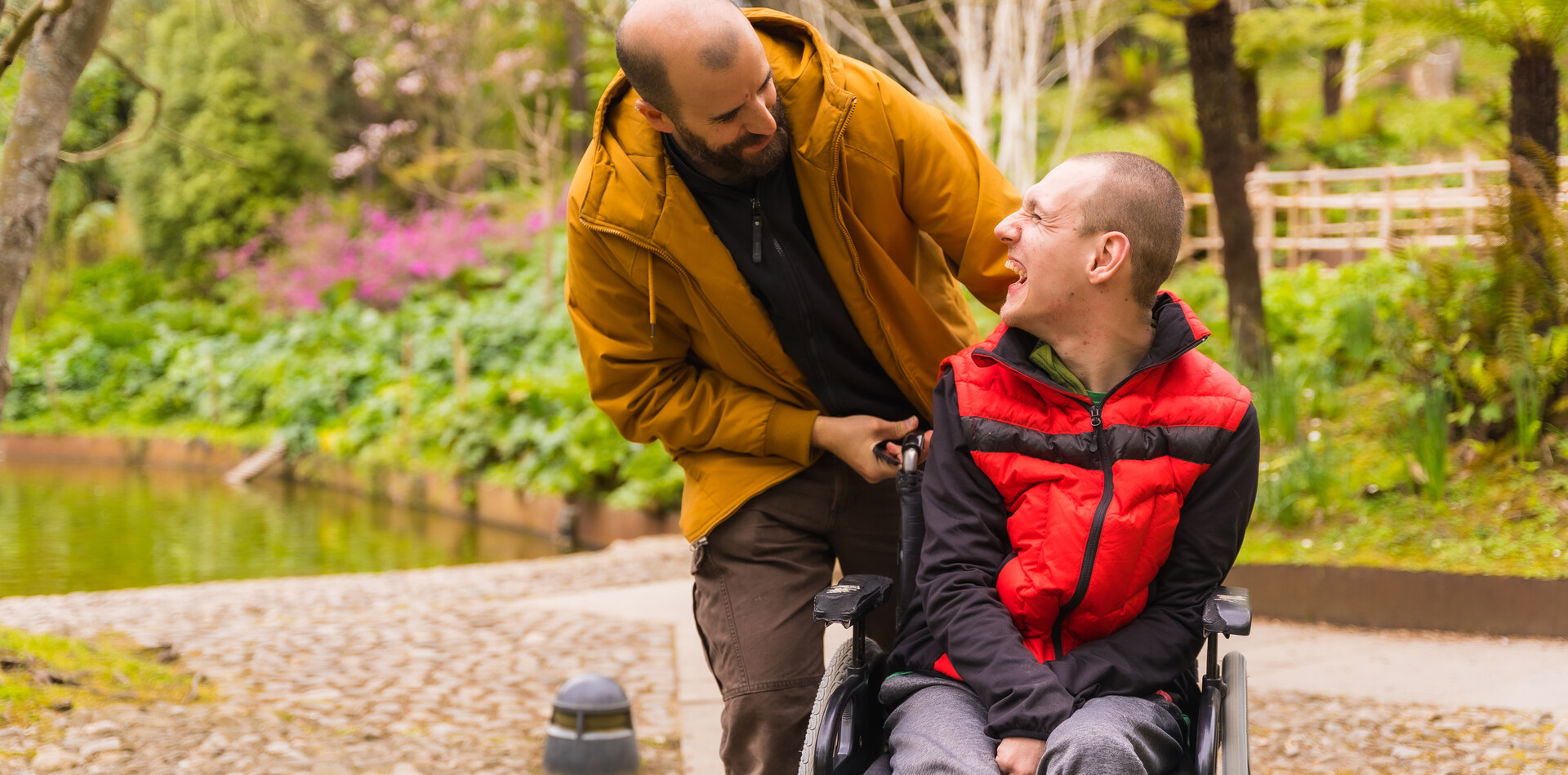 Ein Mann mit gelber Jacke schiebt einen anderen Mann im Rollstuhl, er neigt sich nach vorne und sie schauen einander lachend an. Im Hintergrund sind B&auml;ume und Str&auml;ucher.