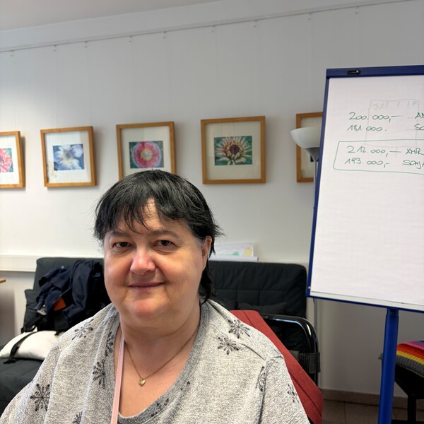 Portr&auml;t einer leicht l&auml;chelnden Frau mit kurzen, dunklen Haaren (Helga), die ein graues Oberteil mit schwarzen Schneeflocken-Mustern und eine feine Halskette tr&auml;gt. Sie sitzt in einem hellen Raum; im Hintergrund h&auml;ngen gerahmte Bilder an der Wand und rechts steht ein Flipchart mit handschriftlichen Notizen.
