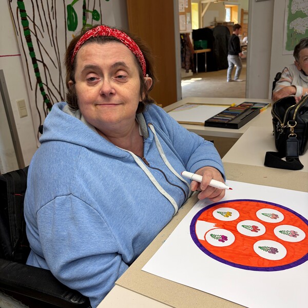 Die K&uuml;nstlerin Gabi M&uuml;hlbauer mit einem roten Haarband und einem hellblauen Kapuzenpullover sitzt l&auml;chelnd an einem Tisch und zeichnet. Vor ihr liegt ein wei&szlig;es Blatt Papier, auf dem ein rundes, farbenfrohes Kunstwerk mit Blumenmotiven in Rot- und Lilat&ouml;nen zu sehen ist. Sie h&auml;lt einen wei&szlig;en Filzstift in der Hand; im Hintergrund sind ein Malkasten und weitere Zeichnungen erkennbar.