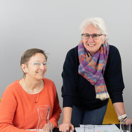 Die Ansprechpersonen der BALANCE-Telefonzentrale an einem Arbeitstisch vor hellem Hintergrund. Links sitzt Barbara Toth in einem leuchtend orangefarbenen Oberteil und blickt l&auml;chelnd in die Kamera. In der Mitte steht Martina Steinh&auml;user, die sich mit den H&auml;nden auf den Tisch st&uuml;tzt. Sie tr&auml;gt einen dunkelblauen Pullover und einen bunt gemusterten Schal in Violett- und Oranget&ouml;nen. Vor ihnen auf dem Tisch liegen Arbeitsunterlagen und Wassergl&auml;ser.