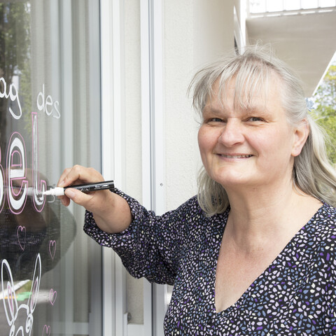 Eine Teilnehmerin der Tagesstruktur K&auml;the Dorsch steht l&auml;chelnd vor einer Glasscheibe und schreibt mit einem wei&szlig;en Kreidemarker die Worte &sbquo;Welttag des Esels&lsquo; auf das Fenster. Unter dem Schriftzug sind eine kleine Esel-Illustration und Herzen zu sehen. Die Frau mit grauem Haar tr&auml;gt eine dunkle Bluse mit feinem Muster und blickt freundlich in die Kamera.