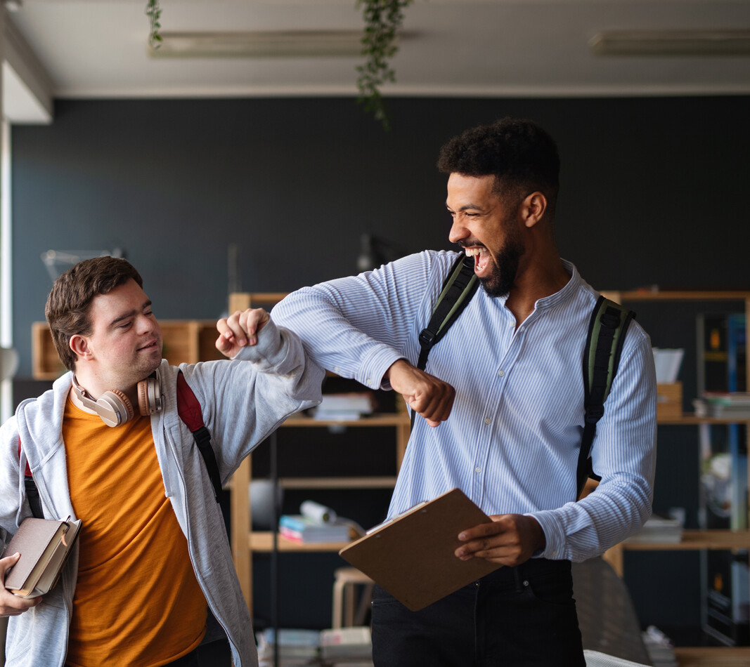 Zwei junge M&auml;nner begr&uuml;&szlig;en sich in einem hellen, modernen Innenraum mit einem freundschaftlichen Ellenbogencheck. Beide tragen Rucks&auml;cke und halten B&uuml;cher bzw. ein Klemmbrett in den H&auml;nden. Der linke Mann mit Trisomie 21 l&auml;chelt leicht, w&auml;hrend der rechte Mann mit dunkleren Locken und Bart herzlich lacht. Die Szene vermittelt eine Atmosph&auml;re von Freude, Gemeinschaft und gelebter Inklusion.