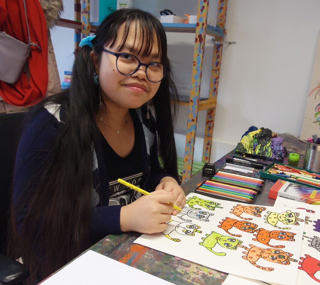 Die K&uuml;nstlerin Ashley Horney sitzt an ihrem Arbeitsplatz und zeichnet auf ein Blatt Papier. Sie l&auml;chelt in die Kamera. Sie hat lange dunkle Haare, tr&auml;gt eine Brille und ein dunkelblaues Shirt mit wei&szlig;em Schriftaufdruck.