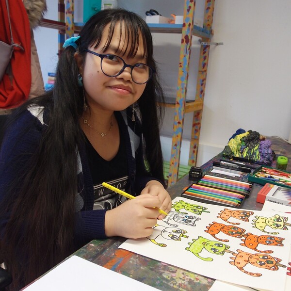Die K&uuml;nstlerin Ashley Horney sitzt an ihrem Arbeitsplatz und zeichnet auf ein Blatt Papier. Sie l&auml;chelt in die Kamera. Sie hat lange dunkle Haare, tr&auml;gt eine Brille und ein dunkelblaues Shirt mit wei&szlig;em Schriftaufdruck.