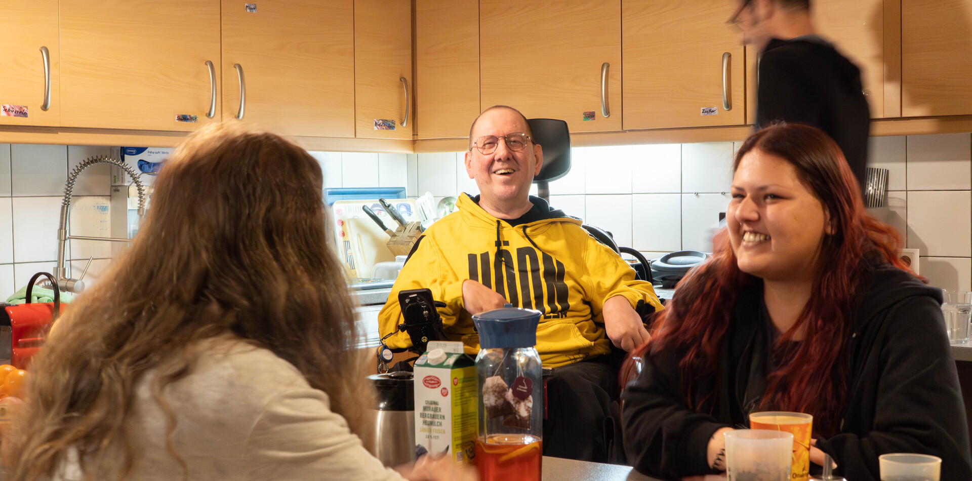 Drei Menschen sitzen gemeinsam an einem K&uuml;chentisch und unterhalten sich lachend; im Hintergrund ist eine K&uuml;chenzeile zu sehen.