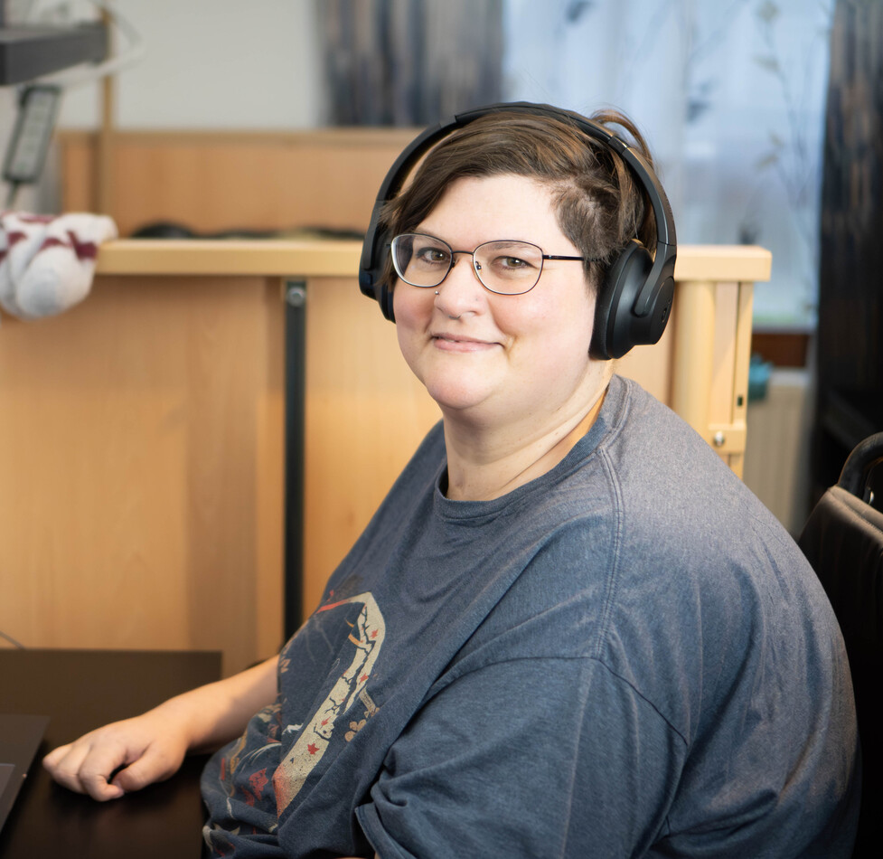 Person mit Kopfh&ouml;rern sitzt an einem Schreibtisch und l&auml;chelt in die Kamera &ndash; eine Alltagssituation beim selbstst&auml;ndigen Wohnen.