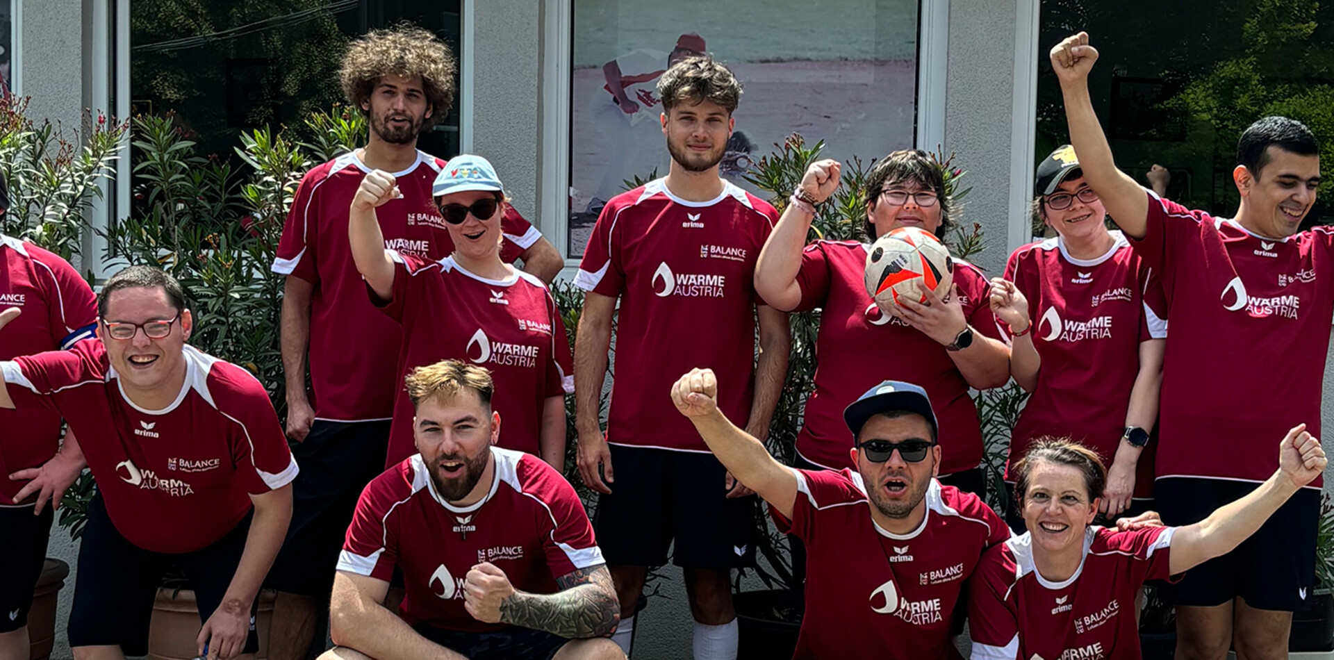 Das Fu&szlig;ballteam des Vereins BALANCE posiert jubelnd f&uuml;r ein Gruppenfoto vor einem Geb&auml;ude. Die elf Spielerinnen und Spieler tragen weinrote Trikots mit den Logos von BALANCE und &bdquo;W&auml;rme Austria&ldquo;. Einige Teammitglieder hocken in der vorderen Reihe, andere stehen dahinter, recken die F&auml;uste in die Luft oder halten einen Fu&szlig;ball in der Hand.