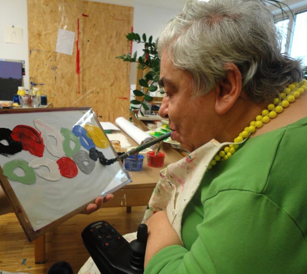 Die K&uuml;nstlerin Lisi Hinterlechner malt mit einem Pinsel, den sie mit dem Mund f&uuml;hrt ein vor ihr aufgestelltes Bild. Sie hat kurze graumelierte Haare und tr&auml;gt einen gr&uuml;nen Pullover und eine gelbe Perlenkette.