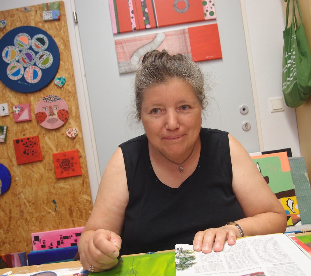 Die K&uuml;nstlerin Bettina Onderka sitzt an ihrem Arbeitsplatz im bildBalance Atelier Wien und blickt mit einem sanften L&auml;cheln in die Kamera. Sie tr&auml;gt ein schwarzes, &auml;rmelloses Oberteil und hat ihr graumeliertes Haar hochgesteckt. Vor ihr auf dem Tisch liegt eine aufgeschlagene Zeitschrift und ein gr&uuml;nes Bild, an dem sie gerade arbeitet. Die Wand hinter ihr ist mit vielen ihrer farbenfrohen, quadratischen und runden Kunstwerke auf Holz und Leinwand geschm&uuml;ckt. Rechts im Bild ist ein Regal mit Malutensilien und einer gr&uuml;nen Stofftasche zu sehen.