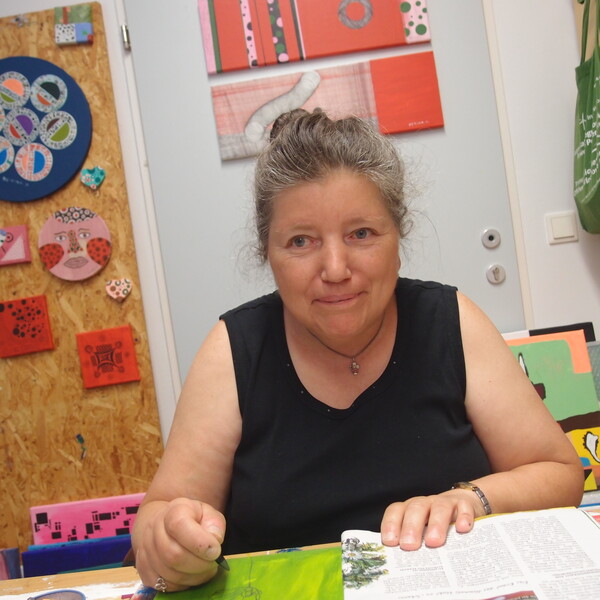 Die K&uuml;nstlerin Bettina Onderka sitzt an ihrem Arbeitsplatz im bildBalance Atelier Wien und blickt mit einem sanften L&auml;cheln in die Kamera. Sie tr&auml;gt ein schwarzes, &auml;rmelloses Oberteil und hat ihr graumeliertes Haar hochgesteckt. Vor ihr auf dem Tisch liegt eine aufgeschlagene Zeitschrift und ein gr&uuml;nes Bild, an dem sie gerade arbeitet. Die Wand hinter ihr ist mit vielen ihrer farbenfrohen, quadratischen und runden Kunstwerke auf Holz und Leinwand geschm&uuml;ckt. Rechts im Bild ist ein Regal mit Malutensilien und einer gr&uuml;nen Stofftasche zu sehen.