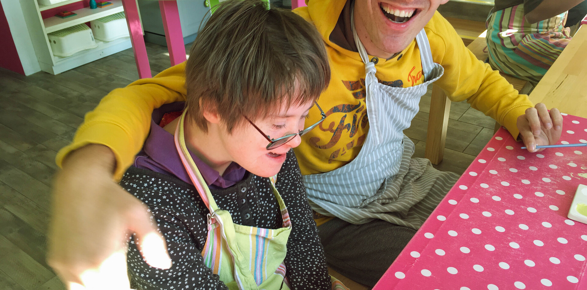 Ein junger Mann und eine junge Frau sitzen an einem Tisch mit einer roten Tischdecke mit weissen Punkten. Sie tragen beide Arbeitssch&uuml;rzen und lachen. Der Mann hat seinne Arm um die Schultern der Frau gelegt. Beide lachen.