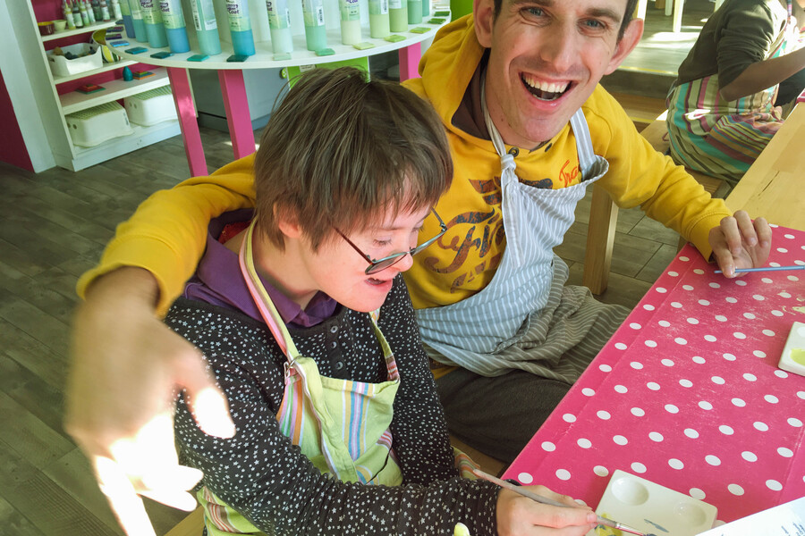 Ein junger Mann und eine junge Frau sitzen an einem Tisch mit einer roten Tischdecke mit weissen Punkten. Sie tragen beide Arbeitssch&uuml;rzen und lachen. Der Mann hat seinne Arm um die Schultern der Frau gelegt. Beide lachen.