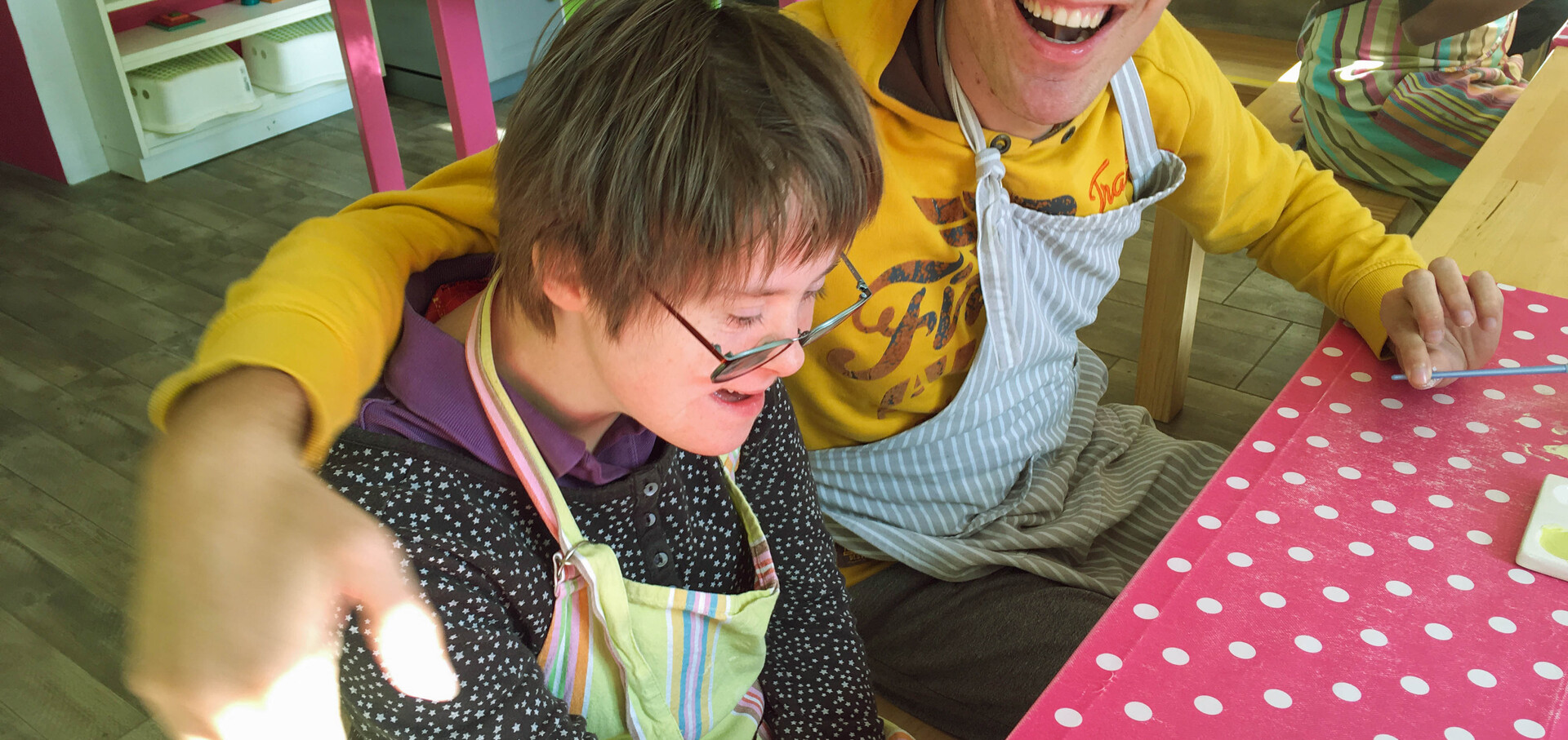 Ein junger Mann und eine junge Frau sitzen an einem Tisch mit einer roten Tischdecke mit weissen Punkten. Sie tragen beide Arbeitssch&uuml;rzen und lachen. Der Mann hat seinne Arm um die Schultern der Frau gelegt. Beide lachen.