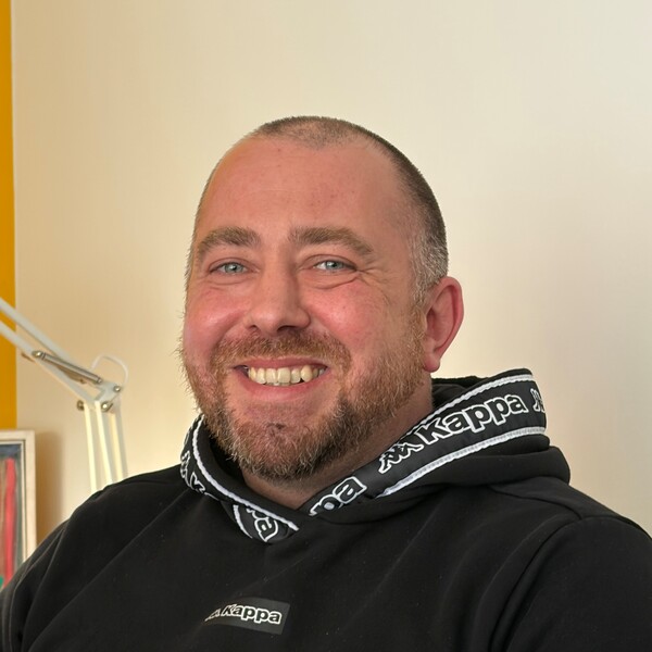 Mitarbeiter Andi sitzt entspannt in einem Innenraum. Er tr&auml;gt einen schwarzen Hoodie mit wei&szlig;en Kappa‑Logos am Kragen und auf der Brust. Im Hintergrund sind eine gelbe Wand, eine Lampe und ein gerahmtes Bild zu sehen.